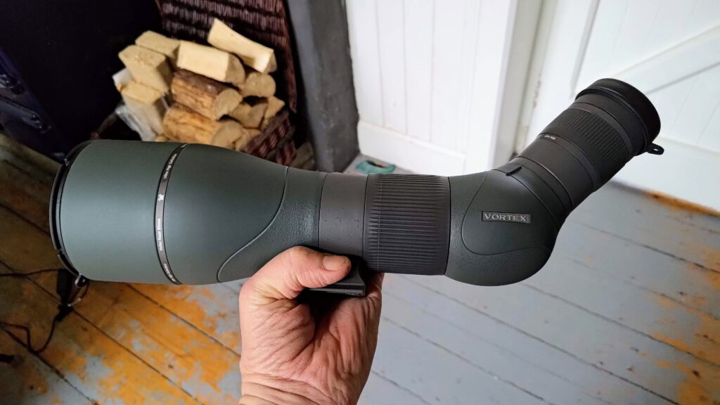A person holding a Vortex Razor HD spotting scope in front of a fireplace, providing a comprehensive review.