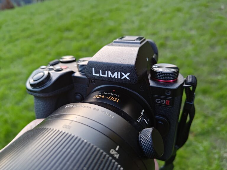 A close-up of a Panasonic Lumix G9II camera held by a hand, with a blurred green grass background.