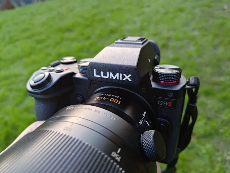 A close-up of a Panasonic Lumix G9II camera held by a hand, with a blurred green grass background.