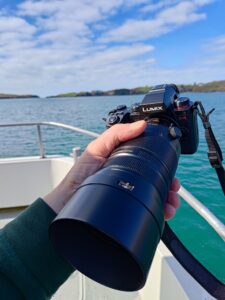 A hand holding a Panasonic Lumix G9II with a large lens on a boat, with a vivid blue sea and green coastline in the background.